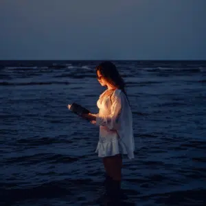 A woman stands in a body of water holding an open book that is illuminating her face and torso.