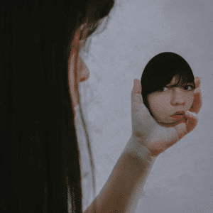 A woman looks into a small handheld mirror, her reflection looking back at the viewer to represent the reflection to be done with this week's astrology.