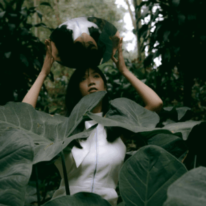 A woman stands amongst large green leaves holding a mirror above her head to represent that reflections to be done using this week's astrology.