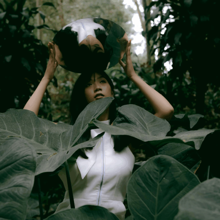 A woman stands amongst large green leaves holding a mirror above her head to represent that reflections to be done using this week's astrology.