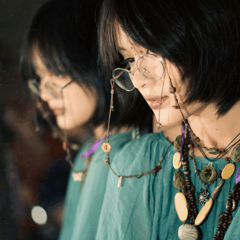 A woman next to a mirror wearing glasses and multiple chunky necklaces. Her reflection is shown in a mirror to represent reflections done during this week's astrology forecast for Nov 10 - 17, 2025.