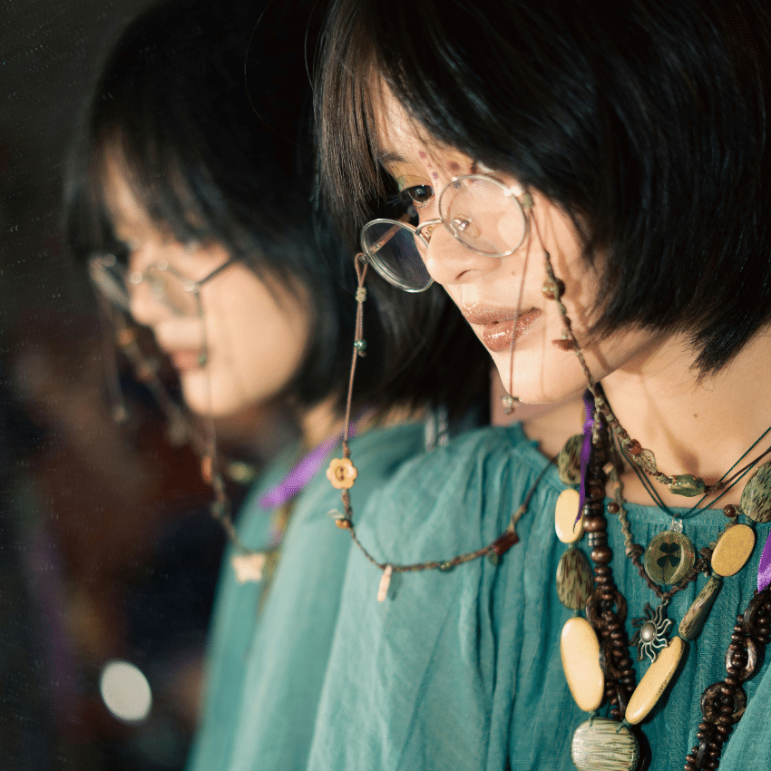 A woman next to a mirror wearing glasses and multiple chunky necklaces. Her reflection is shown in a mirror to represent reflections done during this week's astrology forecast for Nov 10 - 17, 2025.