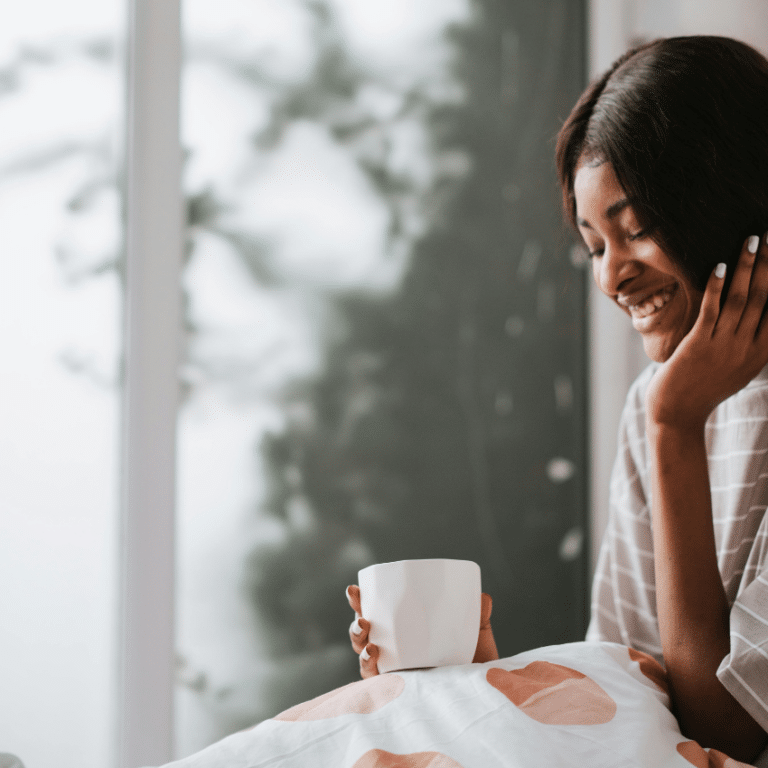 A black woman holds a cup of tea and has a blanket on her lap.