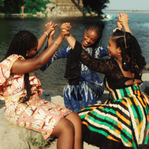Three black woman wearing brightly coloured dresses hold hands, forming a circle.