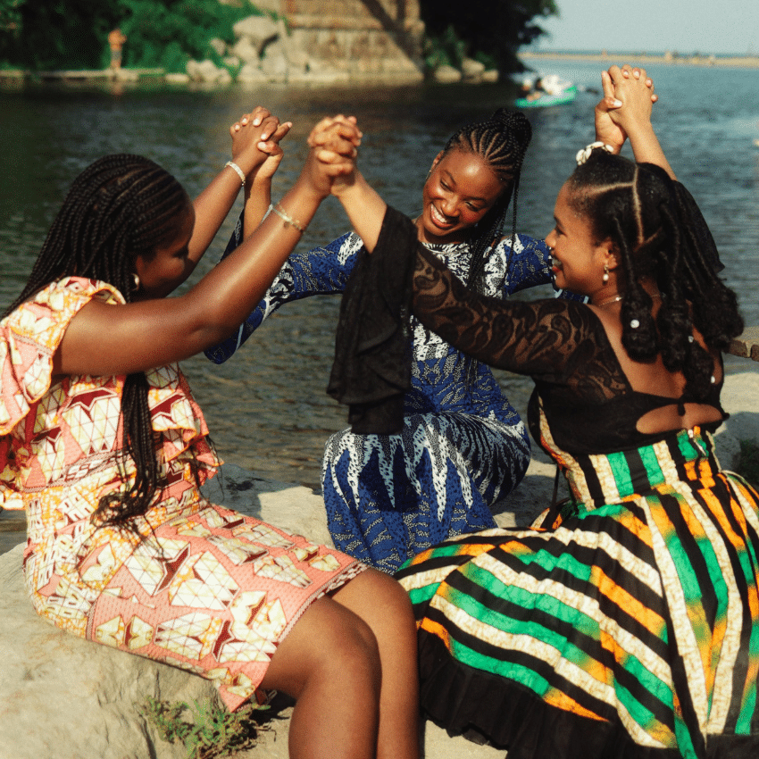 Three black woman wearing brightly coloured dresses hold hands, forming a circle.