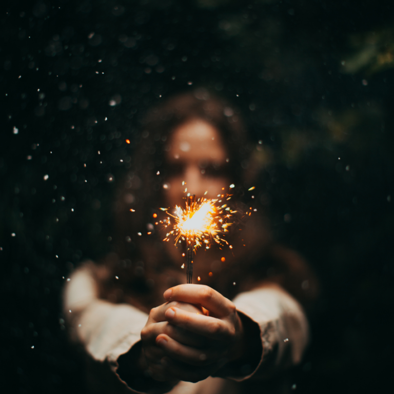 A woman holds a sparkler with two hands in front of her to represent the celebratory energy of January 2026.