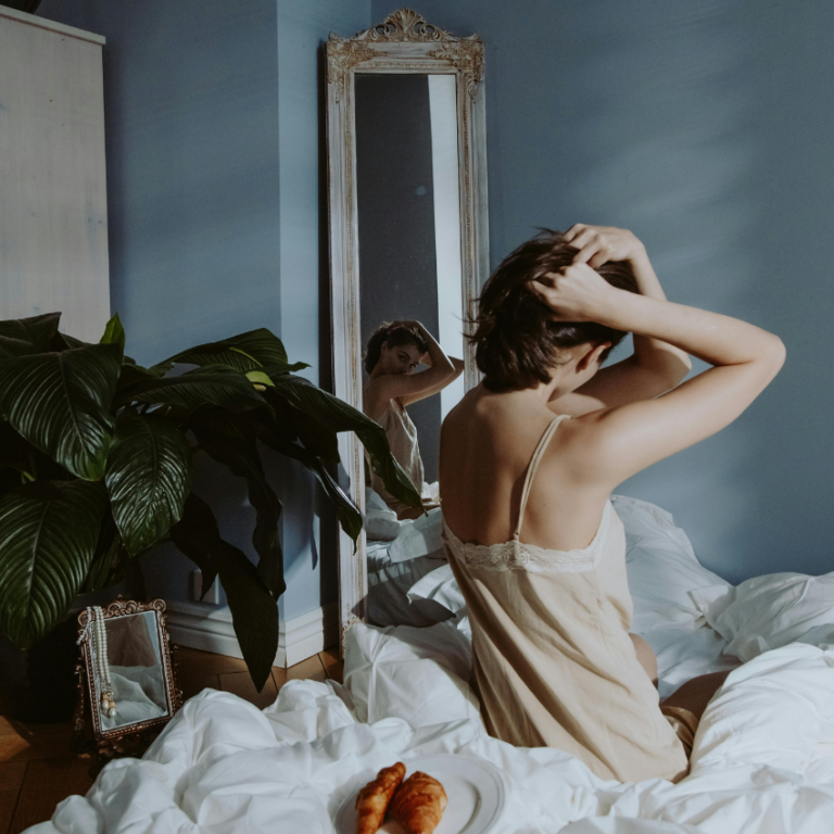 A woman sits on the side of a bed, her reflection shown in an intricate mirror on the wall.