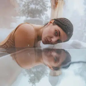 A woman peers into the camera over a frozen pool of water, her reflection visible to represent the weekly astrology reflection.