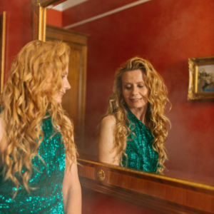 A woman with long, curly red hair looks in a mirror at her fancy green dress to represent the energy of the new year.