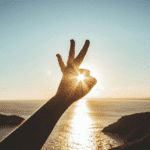 Someone holds their hand up to the sun on the horizon, forming an "ok" sign around it.