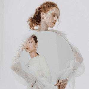 A woman with red hair holds a mirror, reflecting an Asian woman with dark hair. This represents the shift from Capricorn to Aquarius energy during this week's astrology energy.