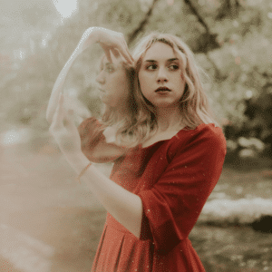 A woman with blonde hair wears a red dress and holds a mirror over her shoulder, her reflection representing the astrological energy of the week.