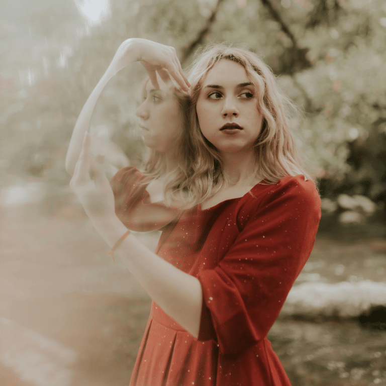 A woman with blonde hair wears a red dress and holds a mirror over her shoulder, her reflection representing the astrological energy of the week.