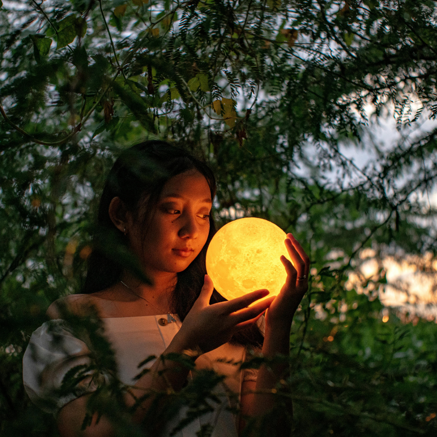 A woman holds a bright orb in front of a tree to represent the upcoming lunar eclipse in Virgo energy.
