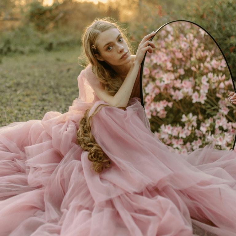 A blonde woman wears a pink gown and holds a mirror that is reflecting pink flowers, representing the spring equinox in March 2026.
