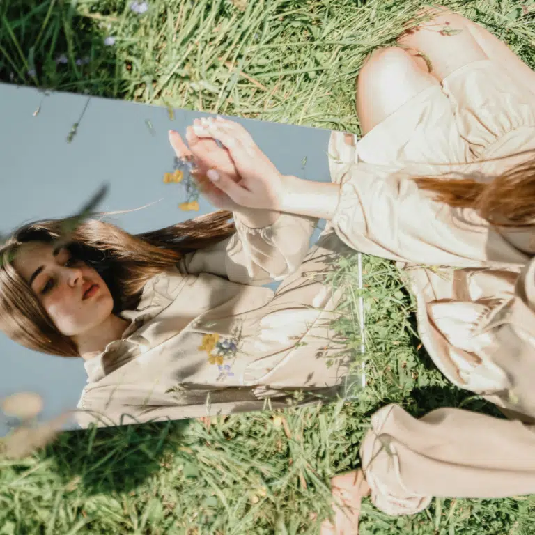 A woman with long brown hair sits on grass and in a looks at a mirror , her downcast reflection representing this weeks astrology reflections.