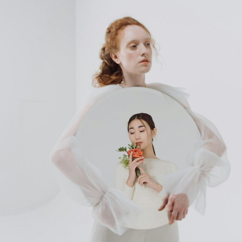A redheaded woman holds a mirror, reflecting an Asian woman holding a flower. The mirror represents the weekly astrology energy and the reflections that need to take place throughout the week.