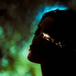A woman is backlit by green and blue light. The only visible part of her face is her eye, representing the energy of the Aries New Moon.