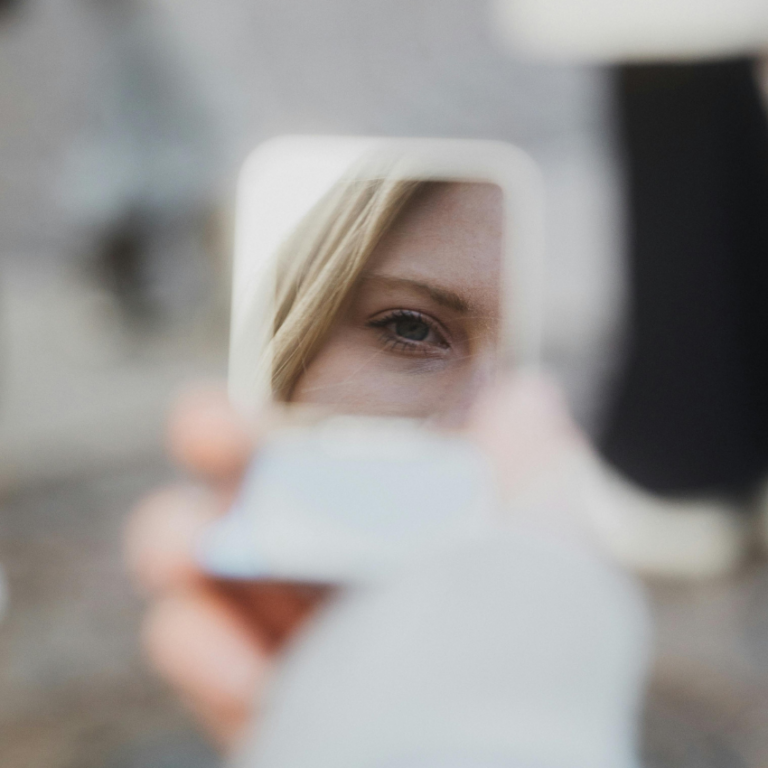 A blonde woman looks into a small square mirror, her reflection the only thing the viewer sees.