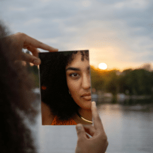 A black woman looks in a handheld mirror, her reflection appearing in front of a body of water and a setting sun, representing this week's astrological energy.