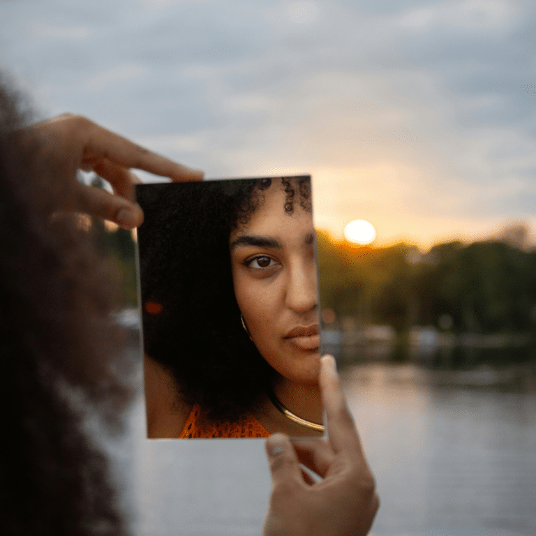 A black woman looks in a handheld mirror, her reflection appearing in front of a body of water and a setting sun, representing this week's astrological energy.