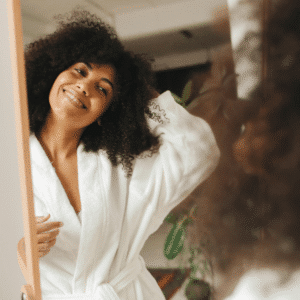 A black woman with a curly afro wearing a white robe looks into a mirror, her smiling reflection representing this week's astrology energy.