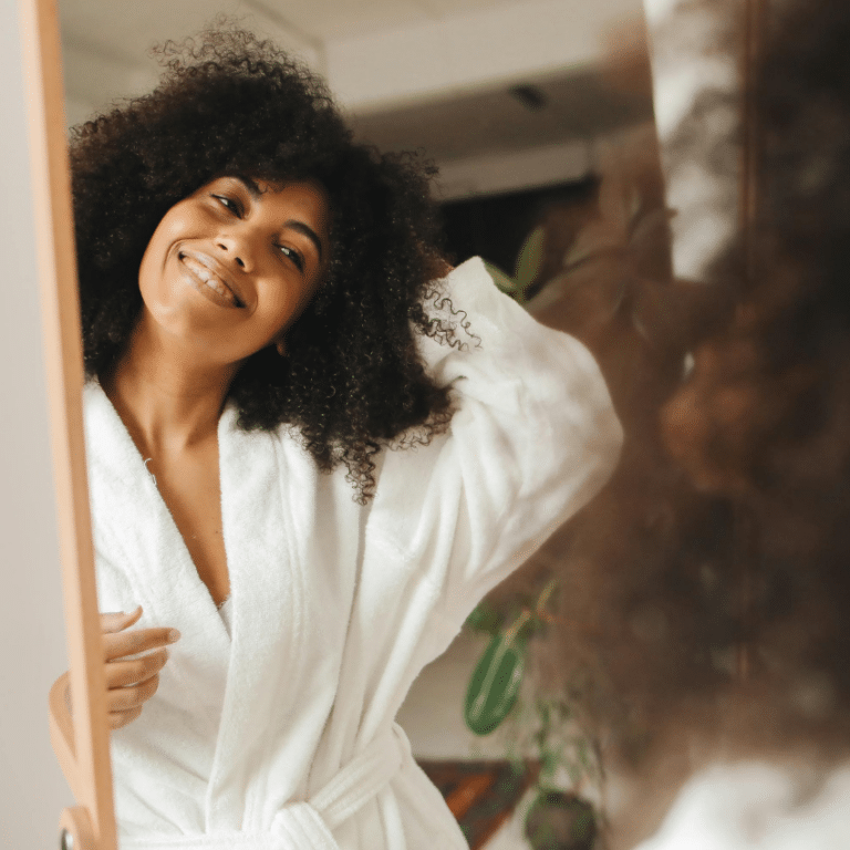 A black woman with a curly afro wearing a white robe looks into a mirror, her smiling reflection representing this week's astrology energy.