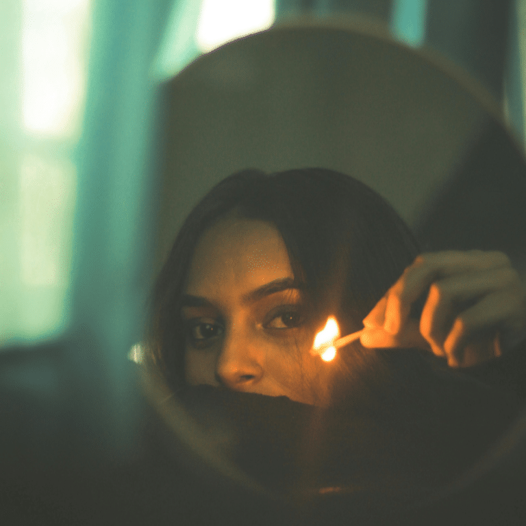A woman holds up a lit match in a mirror, her reflection representing the energy of this week's astrology.