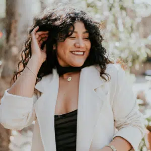 A woman with long brown curly hair turns her head and laughs. There are blossoms on trees in the background, representing the astrological energy of June 2026.