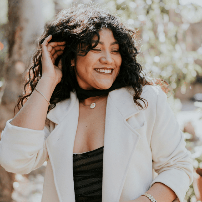 A woman with long brown curly hair turns her head and laughs. There are blossoms on trees in the background, representing the astrological energy of June 2026.