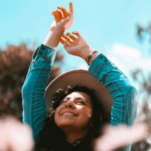 A woman with brown skin and dark hair holds her hands up over her head, appearing to be dancing in abundance.