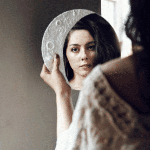 A woman looks in a mirror framed by a Moon decor shape representing the energy of this week's astrology.