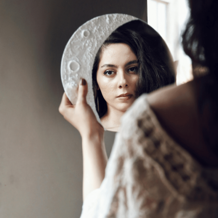 A woman looks in a mirror framed by a Moon decor shape representing the energy of this week's astrology.