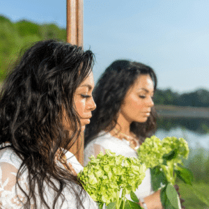 A woman's reflection in a mirror is seen. She is holding green flowers and there is a blue sky in the background.