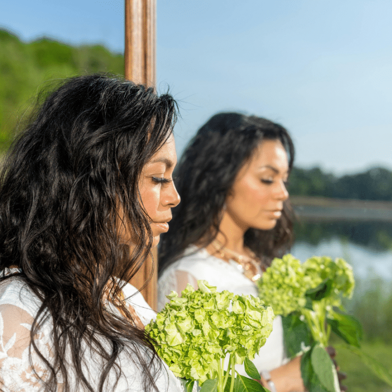 A woman's reflection in a mirror is seen. She is holding green flowers and there is a blue sky in the background.