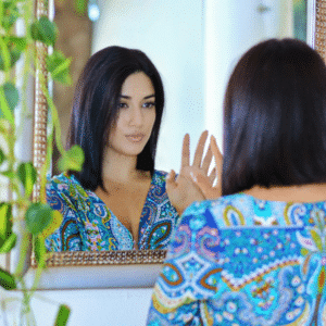 A woman looks in the mirror, touching her hand to her hand's reflection. The mirror is framed on one side with hanging pothos, representing the earthy energy of this week's astrology.