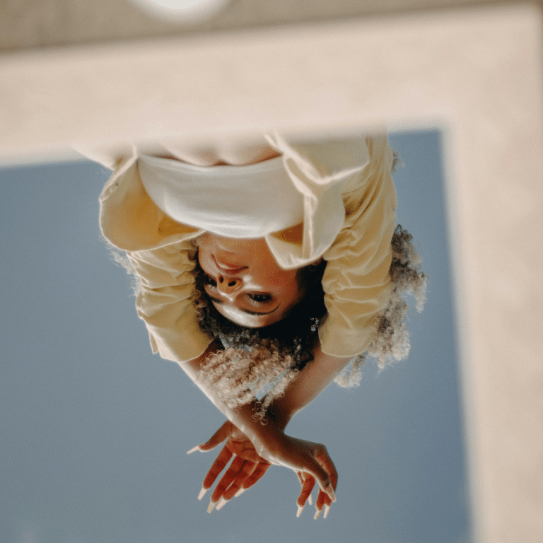 A woman's reflection is seen upside down in a mirror. She is holding her arms up and there is blue sky behind her representing the astrological energy of this week.