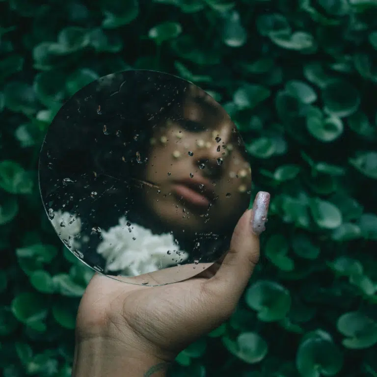 A woman looks in a mirror with a gentle gaze, her reflection in green leaves and a white flower.