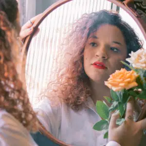 A woman with curly hair holds an orange flower and looks in the mirror, her reflection showing to the viewer.