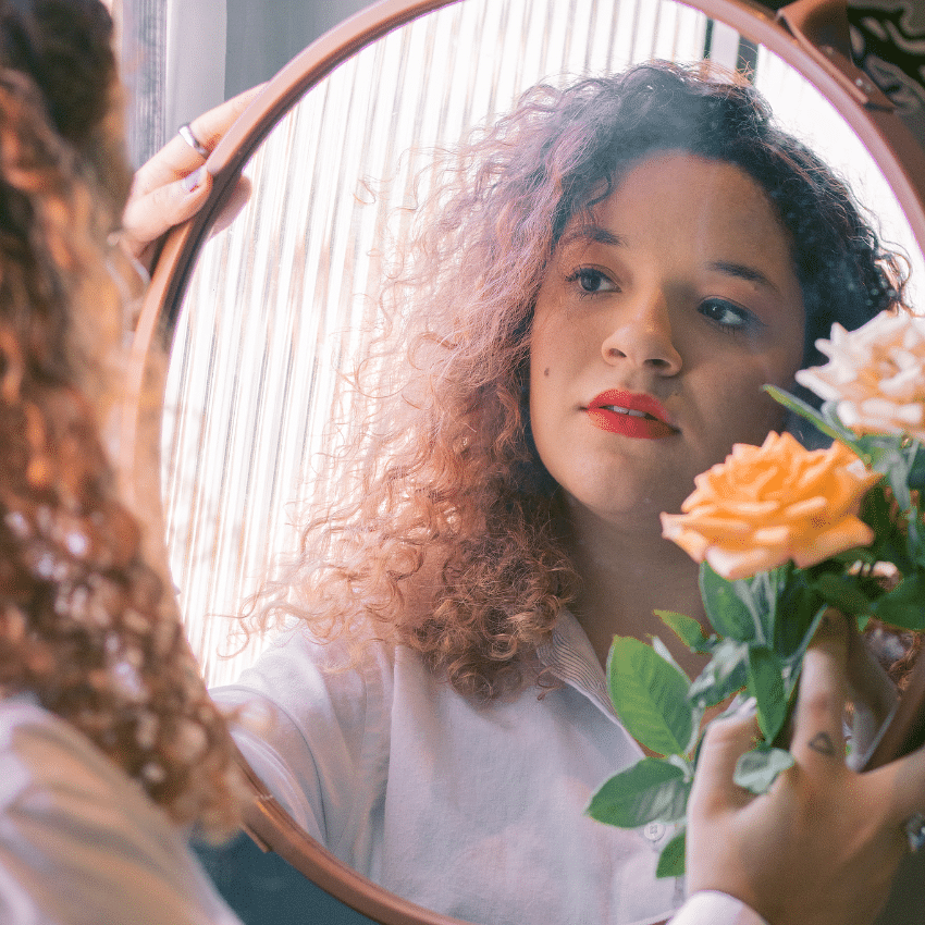 A woman with curly hair holds an orange flower and looks in the mirror, her reflection showing to the viewer.