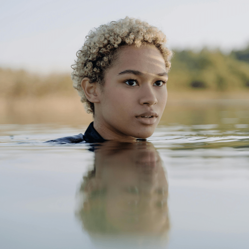 A black woman with short dyed blonde hair emerges from water, her reflection on the surface.