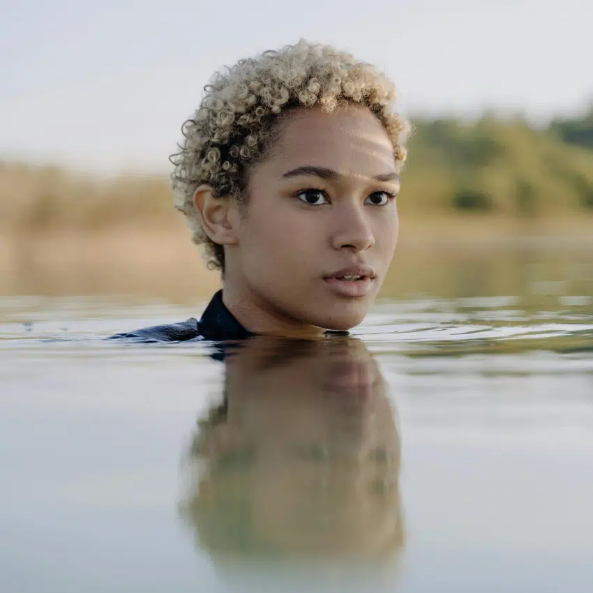 A black woman with short dyed blonde hair emerges from water, her reflection on the surface.