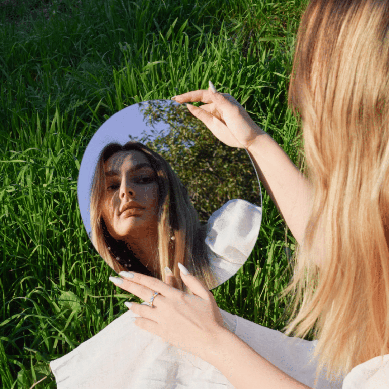 A woman with blonde hair looks in a mirror on a patch of green grass.